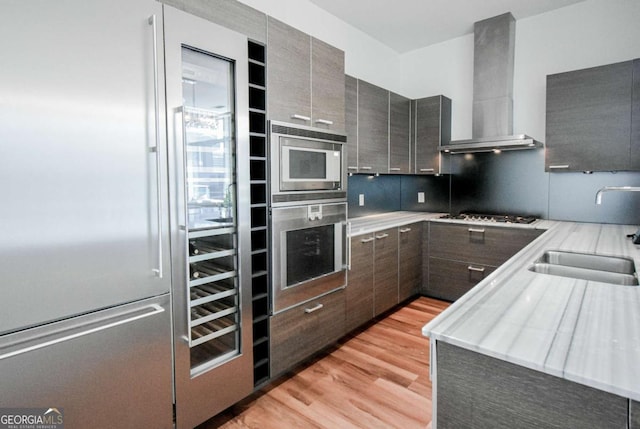 kitchen with wall chimney exhaust hood, modern cabinets, built in appliances, light countertops, and a sink