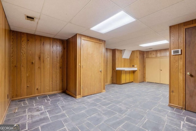 finished basement with a paneled ceiling, wood walls, visible vents, and baseboards