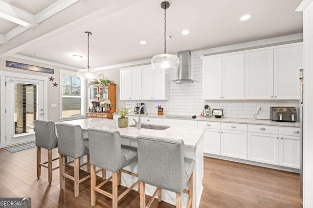 kitchen featuring a center island with sink, wall chimney exhaust hood, light stone counters, white cabinetry, and a sink