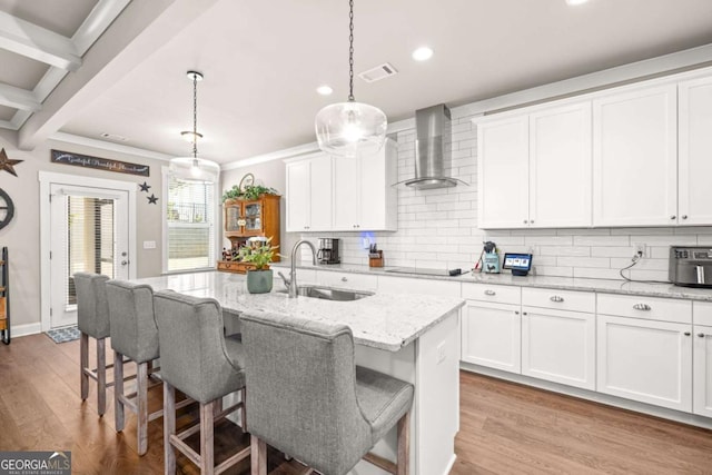 kitchen with light stone counters, pendant lighting, a sink, wall chimney range hood, and an island with sink