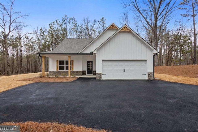 view of front of house with driveway, stone siding, covered porch, a shingled roof, and a garage