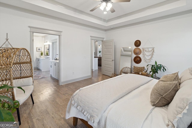 bedroom with a tray ceiling, light wood-style flooring, ceiling fan, ensuite bath, and baseboards