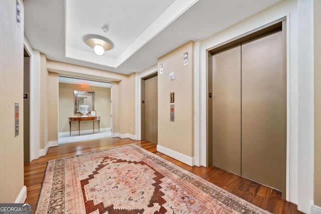 hallway featuring elevator, baseboards, a tray ceiling, and wood finished floors