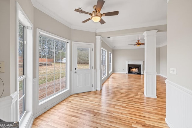 interior space featuring a wainscoted wall, decorative columns, light wood-style flooring, ornamental molding, and a warm lit fireplace