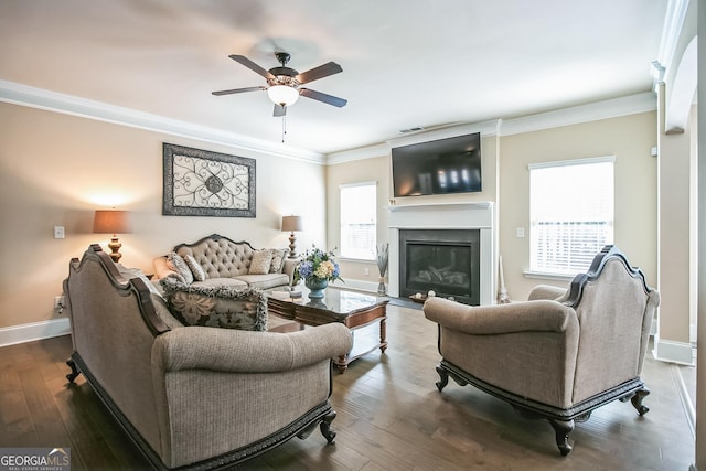 living room featuring baseboards, dark wood finished floors, a glass covered fireplace, ceiling fan, and ornamental molding