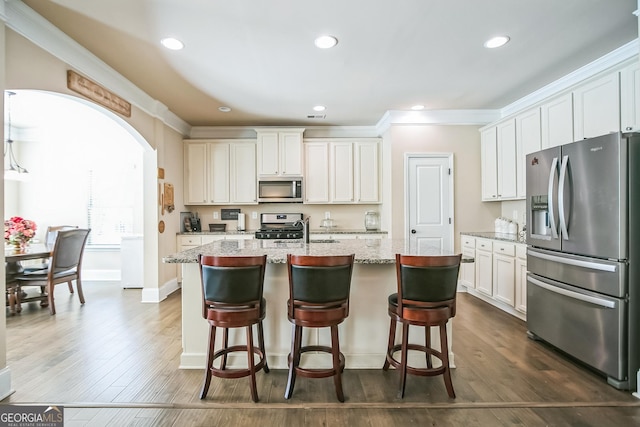 kitchen featuring light stone counters, arched walkways, a center island with sink, stainless steel appliances, and white cabinetry