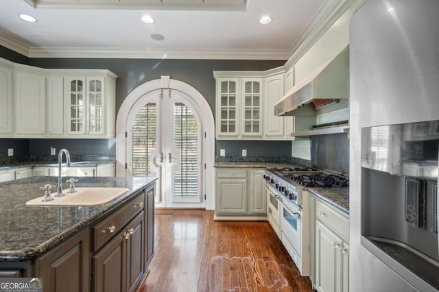 kitchen featuring wall chimney exhaust hood, glass insert cabinets, stainless steel appliances, white cabinetry, and a sink