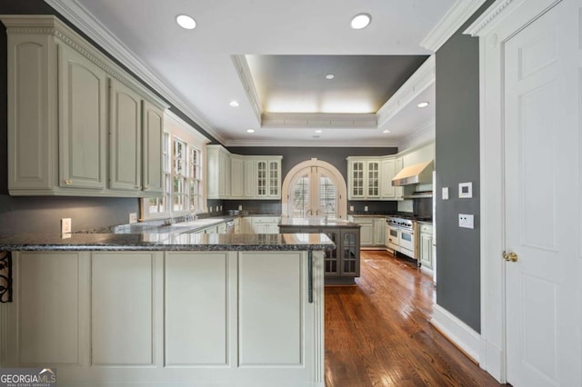 kitchen with a peninsula, double oven range, wall chimney exhaust hood, a raised ceiling, and glass insert cabinets