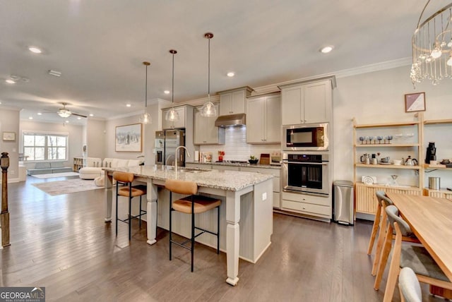 kitchen featuring light stone counters, under cabinet range hood, hanging light fixtures, appliances with stainless steel finishes, and an island with sink