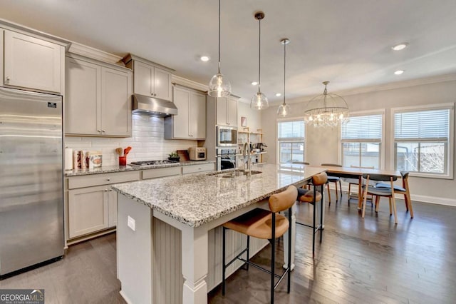 kitchen featuring pendant lighting, an island with sink, light stone countertops, under cabinet range hood, and built in appliances