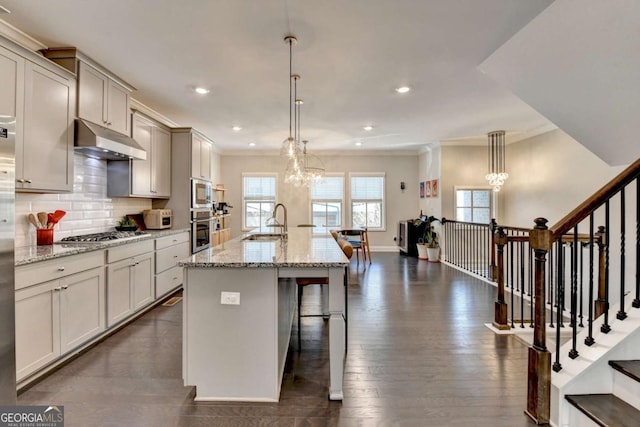 kitchen with under cabinet range hood, light stone countertops, an island with sink, an inviting chandelier, and pendant lighting