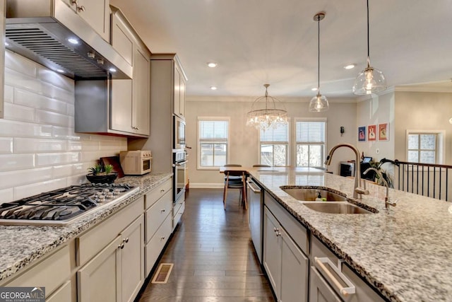 kitchen with light stone counters, stainless steel appliances, a sink, exhaust hood, and hanging light fixtures