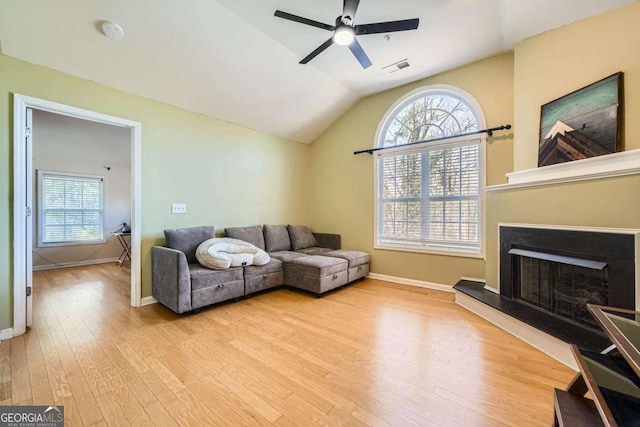 living area featuring a fireplace with raised hearth, lofted ceiling, light wood-style flooring, visible vents, and a ceiling fan