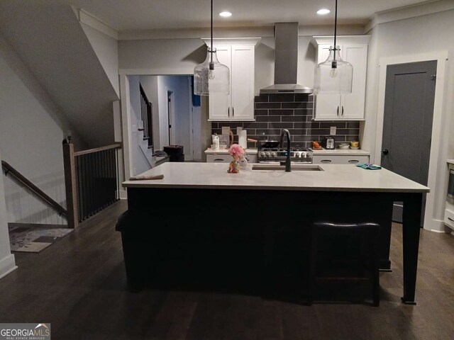 kitchen featuring hanging light fixtures, wall chimney exhaust hood, a center island with sink, and light countertops