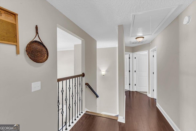 corridor featuring a textured ceiling, dark wood-type flooring, an upstairs landing, baseboards, and attic access