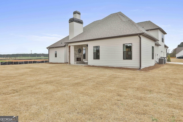 rear view of property with a shingled roof, a chimney, central AC unit, and a lawn