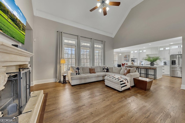 living room with crown molding, a fireplace with raised hearth, wood finished floors, high vaulted ceiling, and baseboards