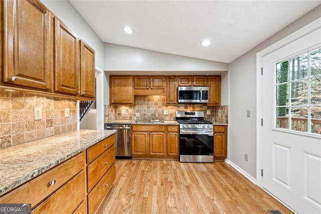 kitchen featuring light stone counters, appliances with stainless steel finishes, brown cabinetry, vaulted ceiling, and a sink