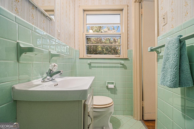 half bathroom featuring a wainscoted wall, tile walls, toilet, vanity, and wallpapered walls