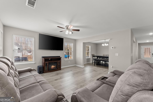 living area featuring light wood finished floors, visible vents, a glass covered fireplace, baseboards, and ceiling fan with notable chandelier