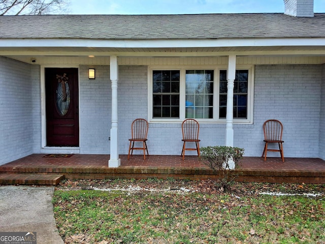 view of exterior entry featuring brick siding, covered porch, and roof with shingles