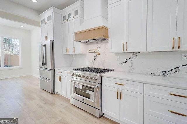 kitchen featuring white cabinets, premium appliances, light stone counters, glass insert cabinets, and premium range hood