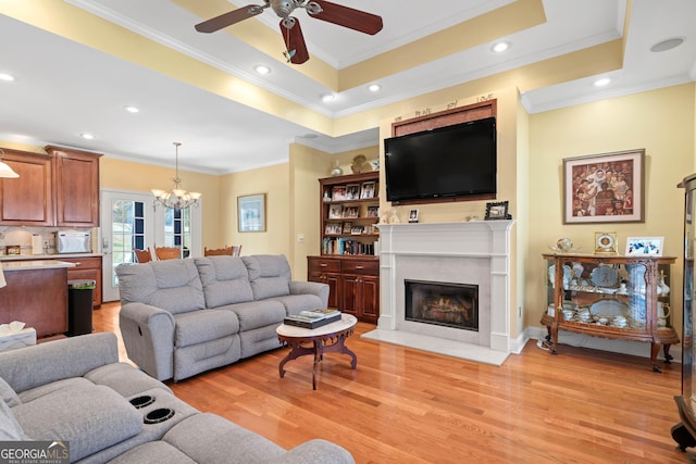 living area with a tray ceiling, crown molding, light wood-style flooring, a fireplace with flush hearth, and ceiling fan with notable chandelier