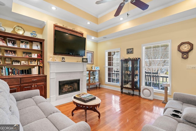 living area featuring a tray ceiling, crown molding, a fireplace with flush hearth, light wood-type flooring, and baseboards