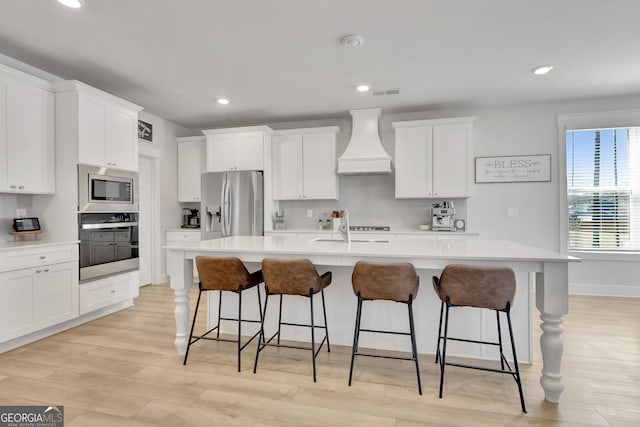 kitchen with an island with sink, custom exhaust hood, stainless steel appliances, and light countertops