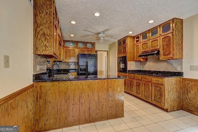 kitchen with under cabinet range hood, a peninsula, brown cabinets, black appliances, and glass insert cabinets