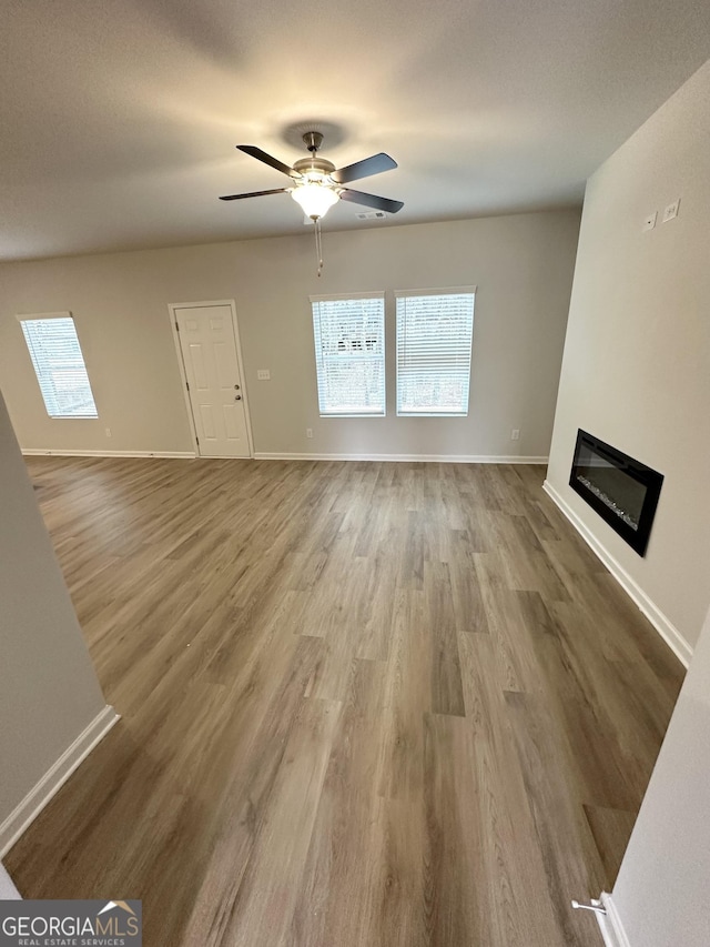 unfurnished living room with a ceiling fan, baseboards, wood finished floors, and a glass covered fireplace