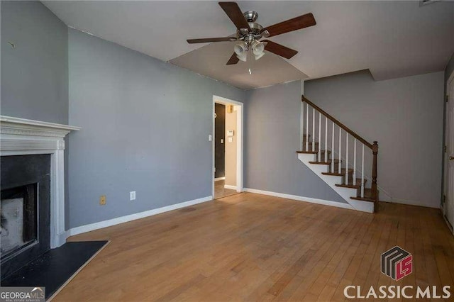 unfurnished living room with a fireplace, stairway, ceiling fan, wood finished floors, and baseboards