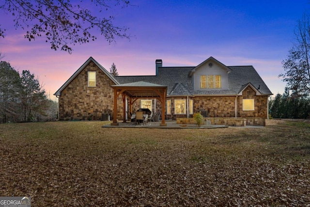 back of property at dusk featuring a patio, stone siding, a chimney, a gazebo, and a yard