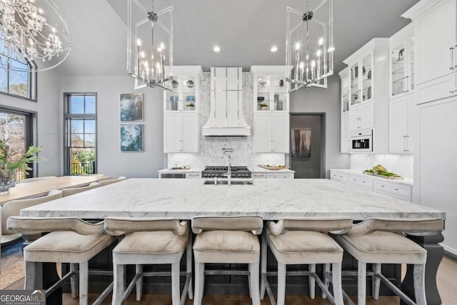 kitchen with glass insert cabinets, white cabinets, custom exhaust hood, a large island with sink, and an inviting chandelier