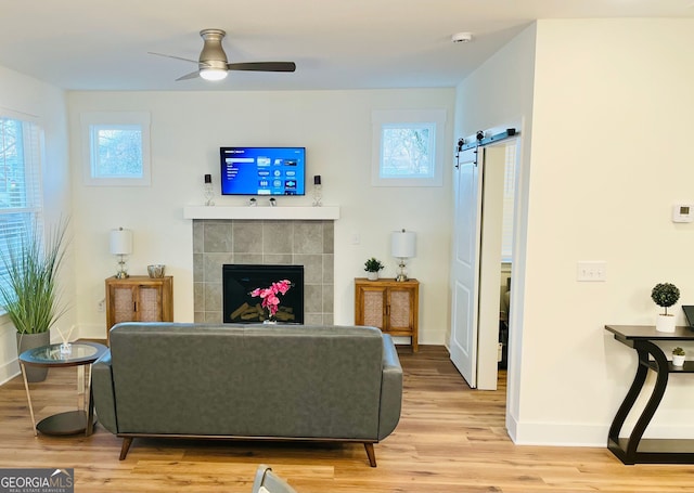 living room featuring light wood-style flooring, a tiled fireplace, a barn door, ceiling fan, and baseboards