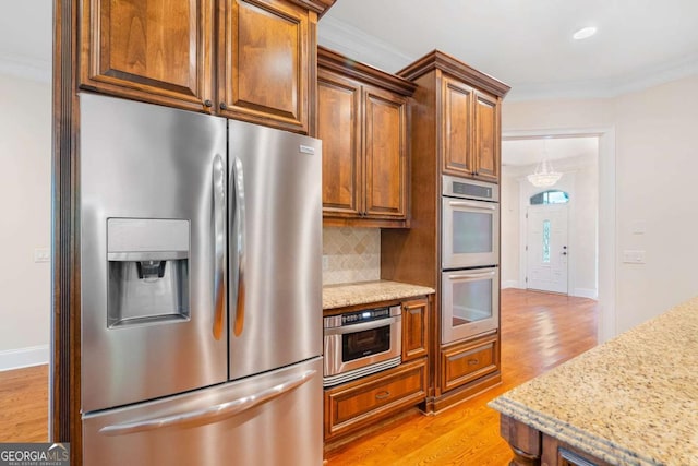 kitchen with light stone counters, backsplash, light wood-style flooring, appliances with stainless steel finishes, and ornamental molding