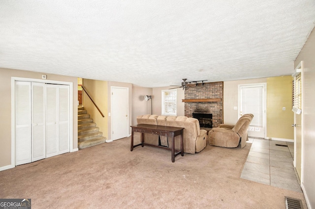 living area featuring ceiling fan, a textured ceiling, visible vents, stairway, and a brick fireplace