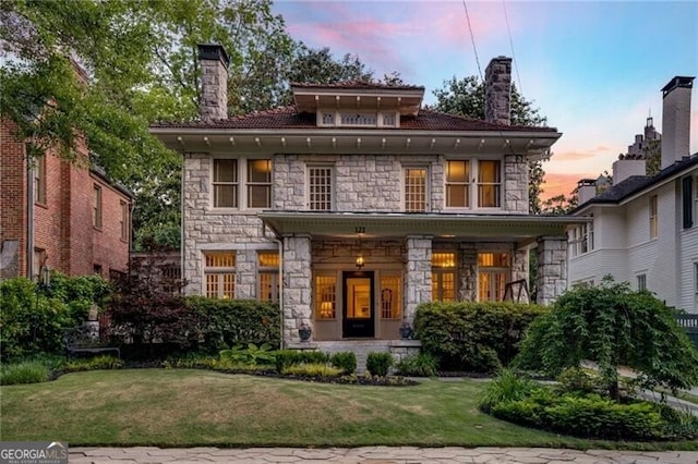 back of property at dusk with stone siding, a lawn, and a chimney