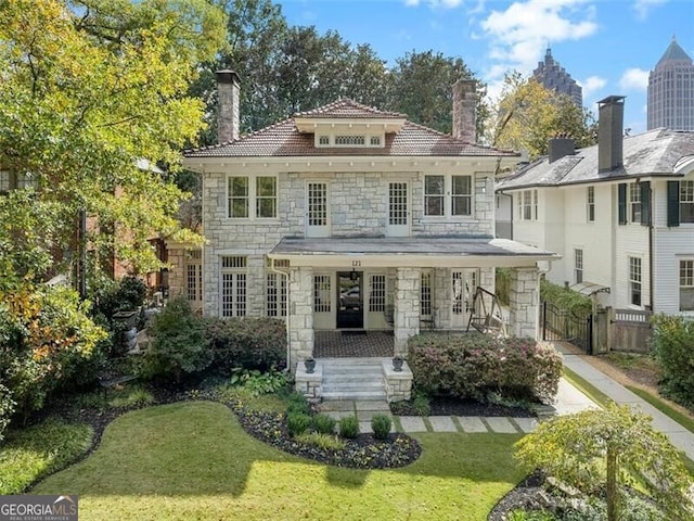 american foursquare style home featuring covered porch, stone siding, a gate, a chimney, and a front yard