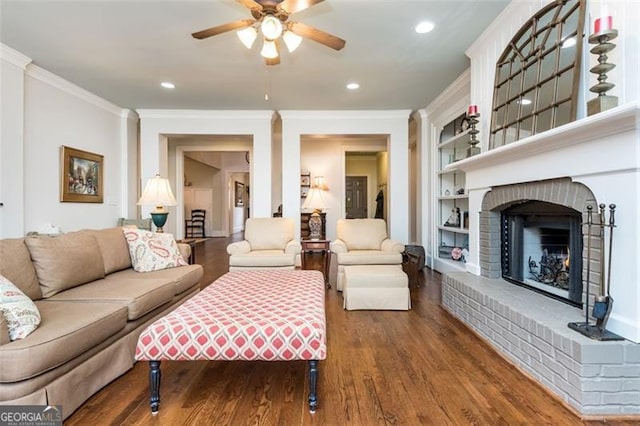 living room with a fireplace, a ceiling fan, crown molding, built in shelves, and wood finished floors