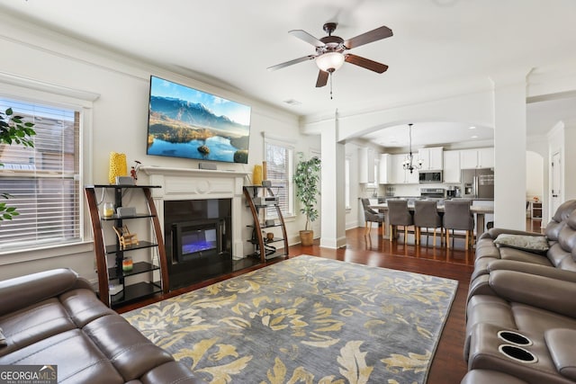 living area featuring visible vents, arched walkways, a glass covered fireplace, dark wood-style flooring, and ceiling fan with notable chandelier