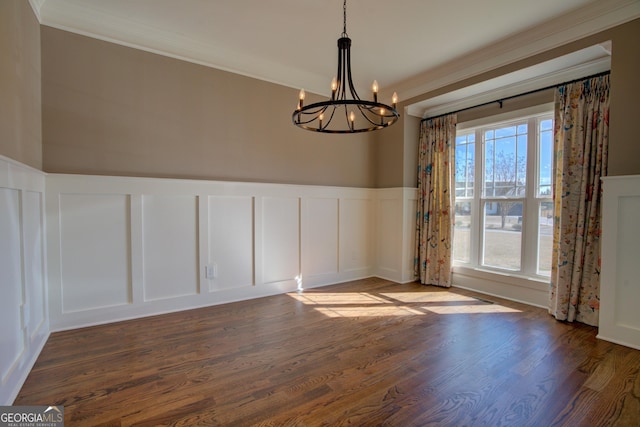 unfurnished dining area with wainscoting, ornamental molding, dark wood-type flooring, an inviting chandelier, and a decorative wall