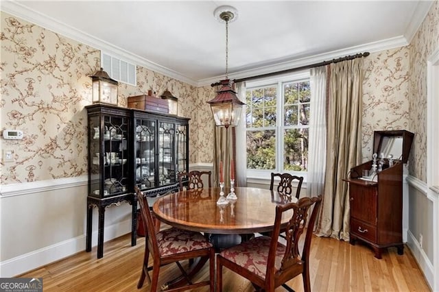 dining room featuring wallpapered walls, light wood finished floors, visible vents, a wainscoted wall, and ornamental molding