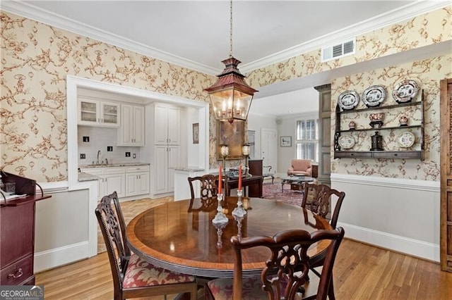 dining room featuring a wainscoted wall, visible vents, ornamental molding, light wood-type flooring, and wallpapered walls