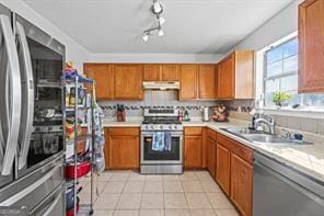 kitchen with under cabinet range hood, light tile patterned floors, brown cabinets, appliances with stainless steel finishes, and a sink