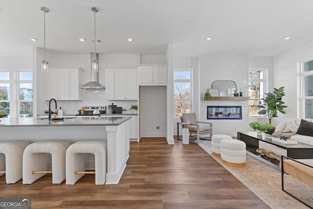 kitchen with white cabinets, an island with sink, hanging light fixtures, wall chimney range hood, and a sink