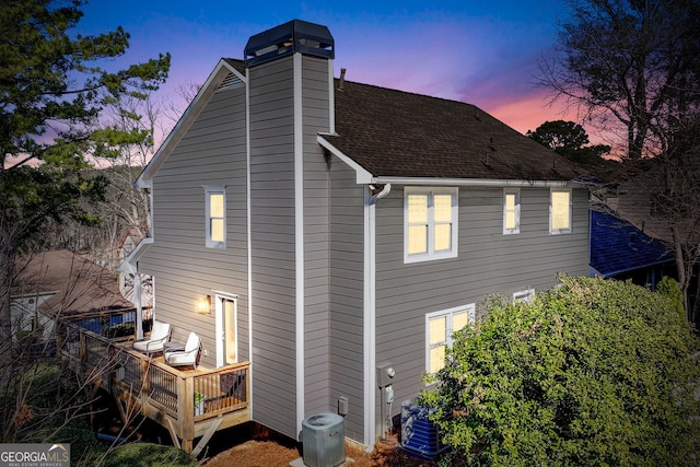 back of property at dusk featuring a shingled roof, a chimney, central AC, and a deck