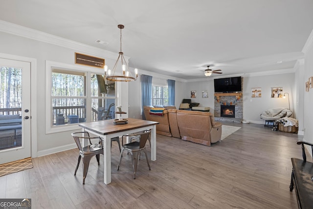 dining room with ceiling fan with notable chandelier, a stone fireplace, wood finished floors, and crown molding