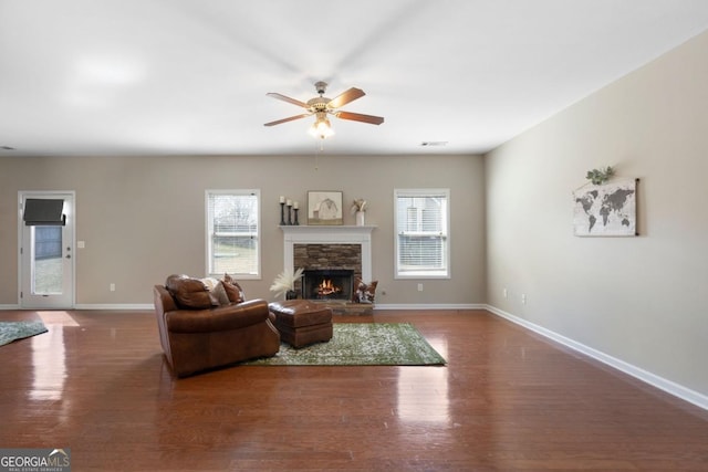 living room with ceiling fan, a stone fireplace, dark wood-type flooring, visible vents, and baseboards