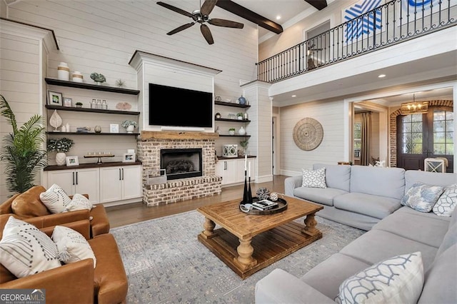 living area featuring ceiling fan with notable chandelier, dark wood-type flooring, a high ceiling, a brick fireplace, and beam ceiling